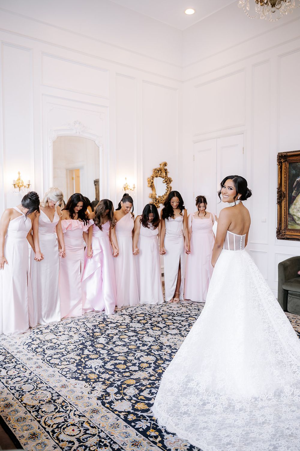 Bride in white ball gown reveals dress to bridesmaids in pink gowns in an elegant white room.