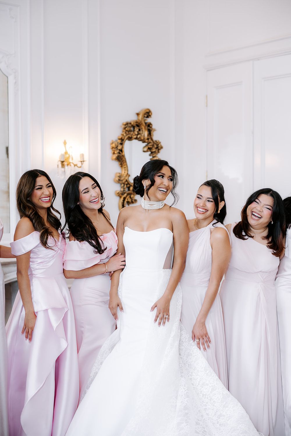 Bride in white gown poses with four bridesmaids in pink dresses, laughing together in an elegant white room.