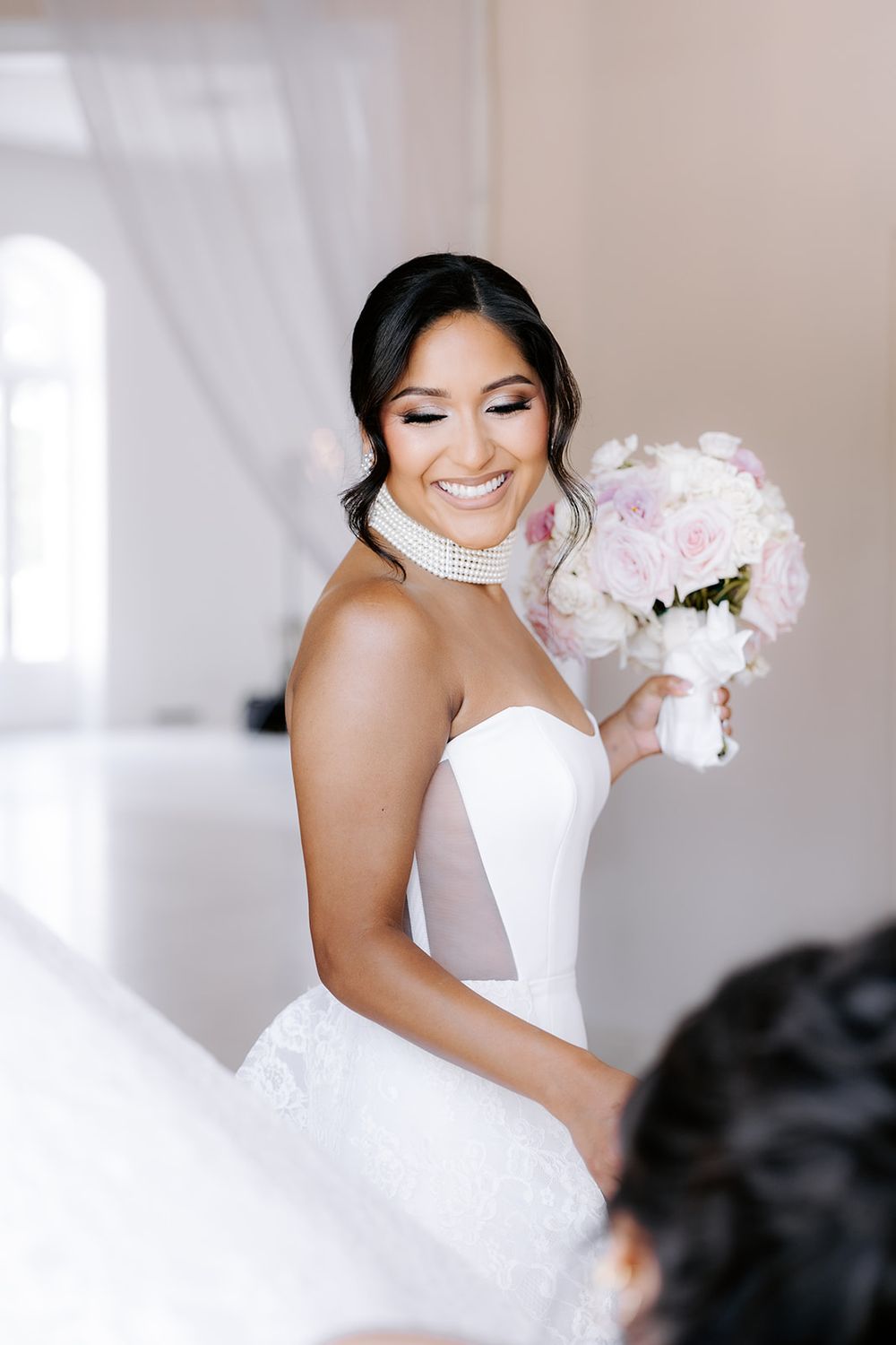 Smiling bride in strapless white gown holds pink and white bouquet, wearing pearl choker necklace in bright elegant room.