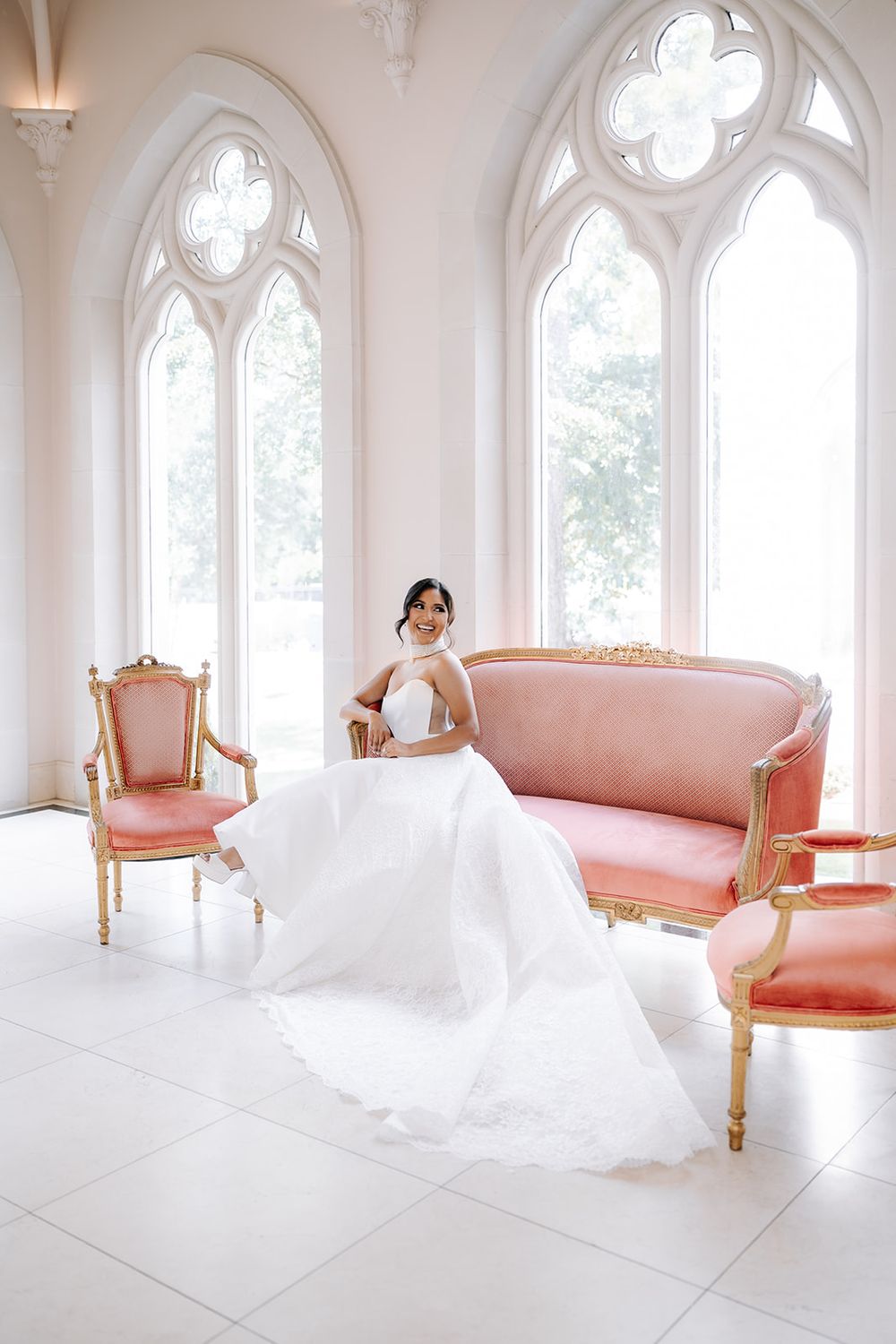 Bride in white gown sits on pink velvet French sofa in bright white chapel with Gothic arched windows.