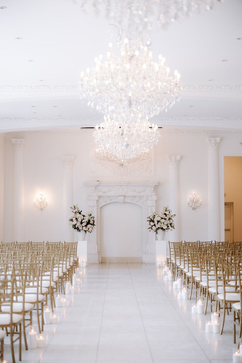 Elegant white wedding ceremony venue with crystal chandelier, gold chiavari chairs, floral arrangements, and candlelit aisle.