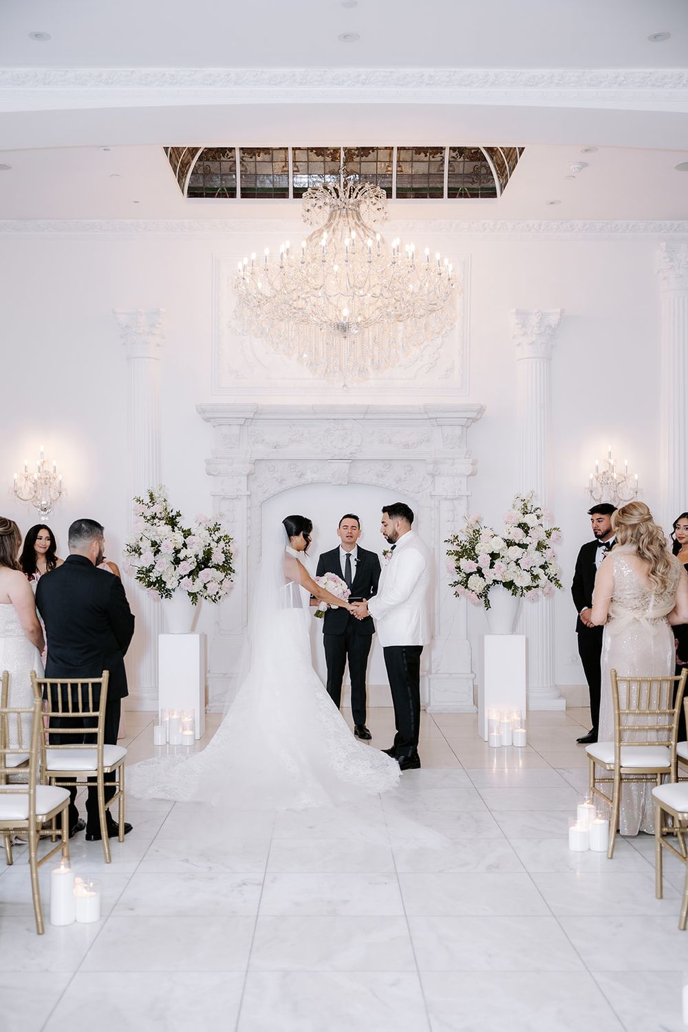 Elegant indoor wedding ceremony with couple at altar, white floral arrangements, crystal chandelier, and marble fireplace backdrop.