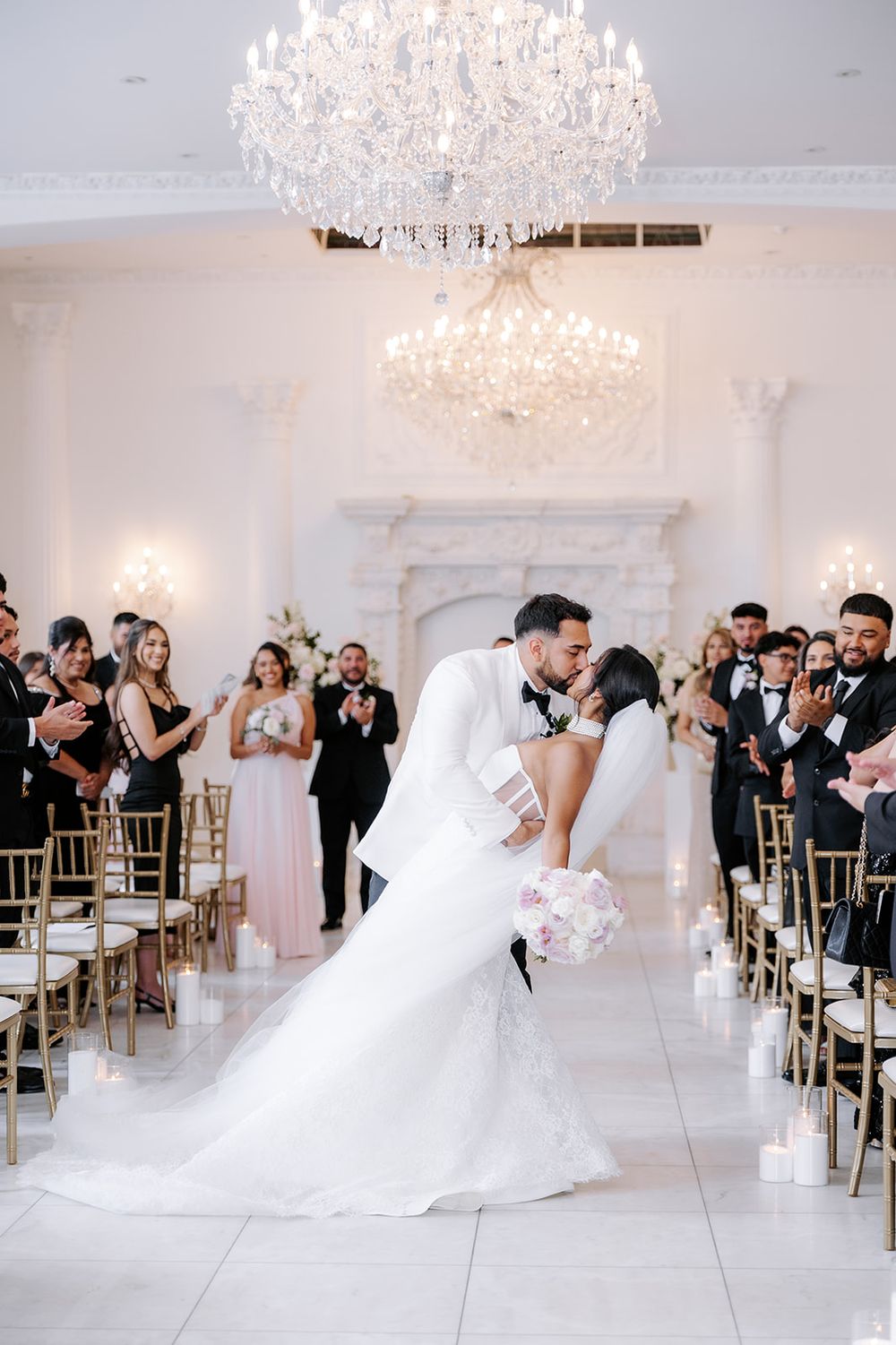 Bride and groom share a romantic dip kiss at the altar in an elegant white ballroom with crystal chandeliers.