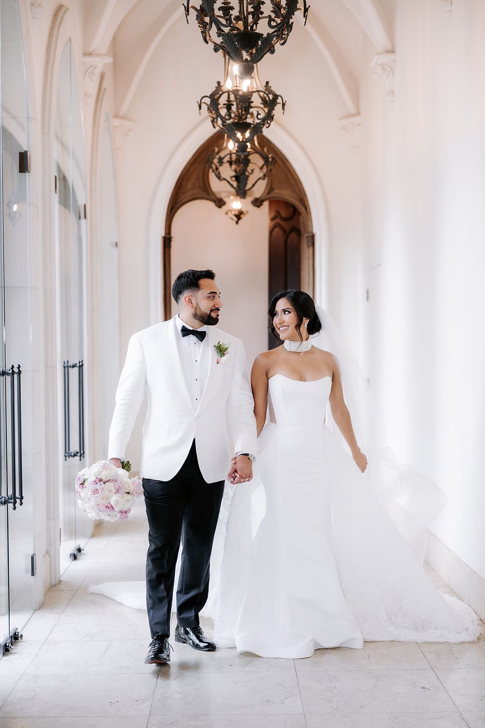 Bride in strapless white gown and groom in white tuxedo jacket hold hands walking through arched venue corridor.