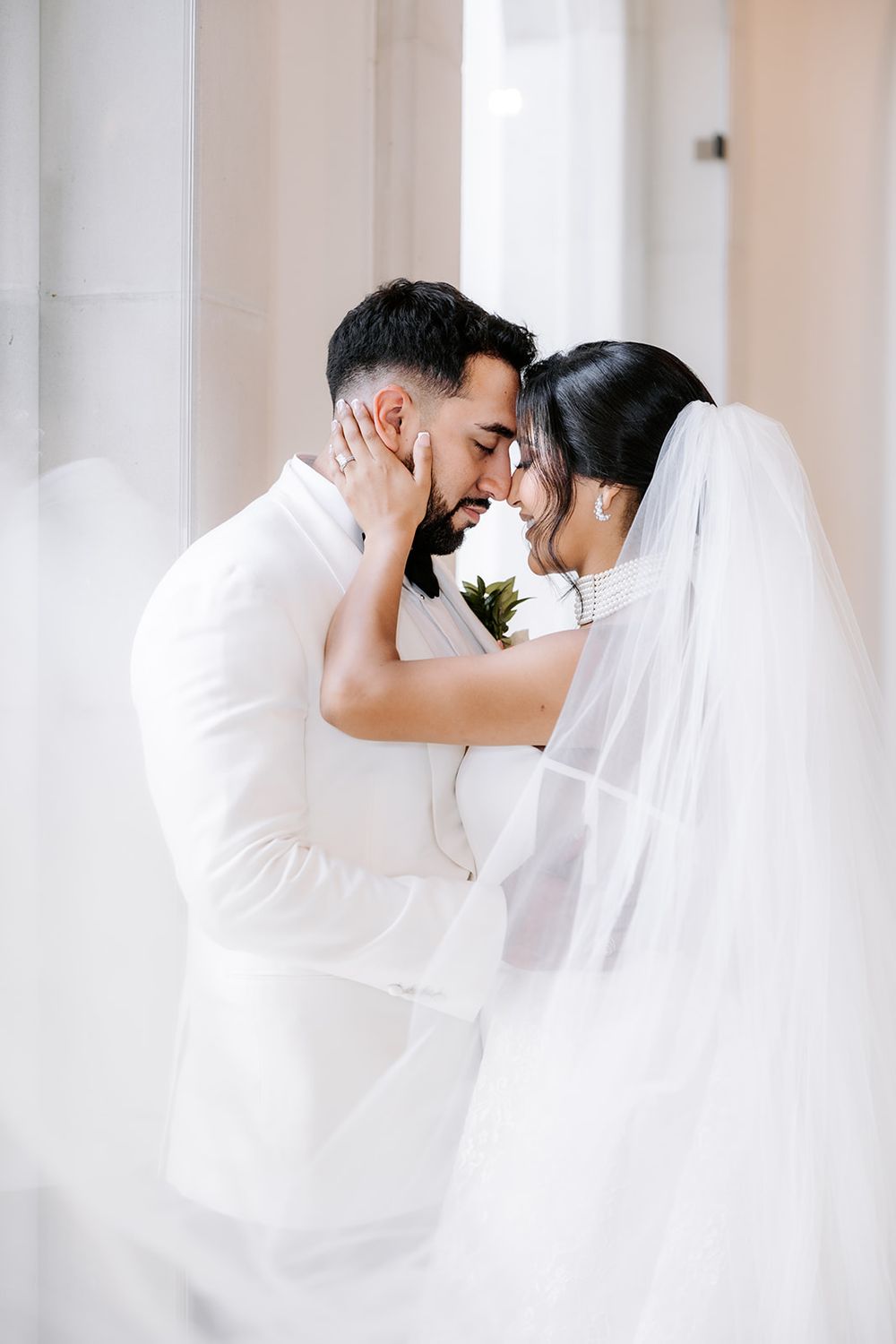 A bride and groom in white attire share a romantic forehead touch, with her veil flowing softly around them.