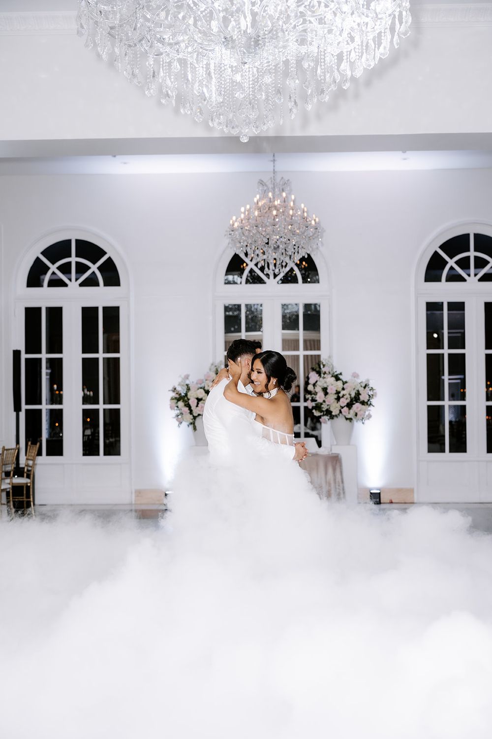 Bride and groom share first dance surrounded by romantic fog under crystal chandeliers in elegant white ballroom.