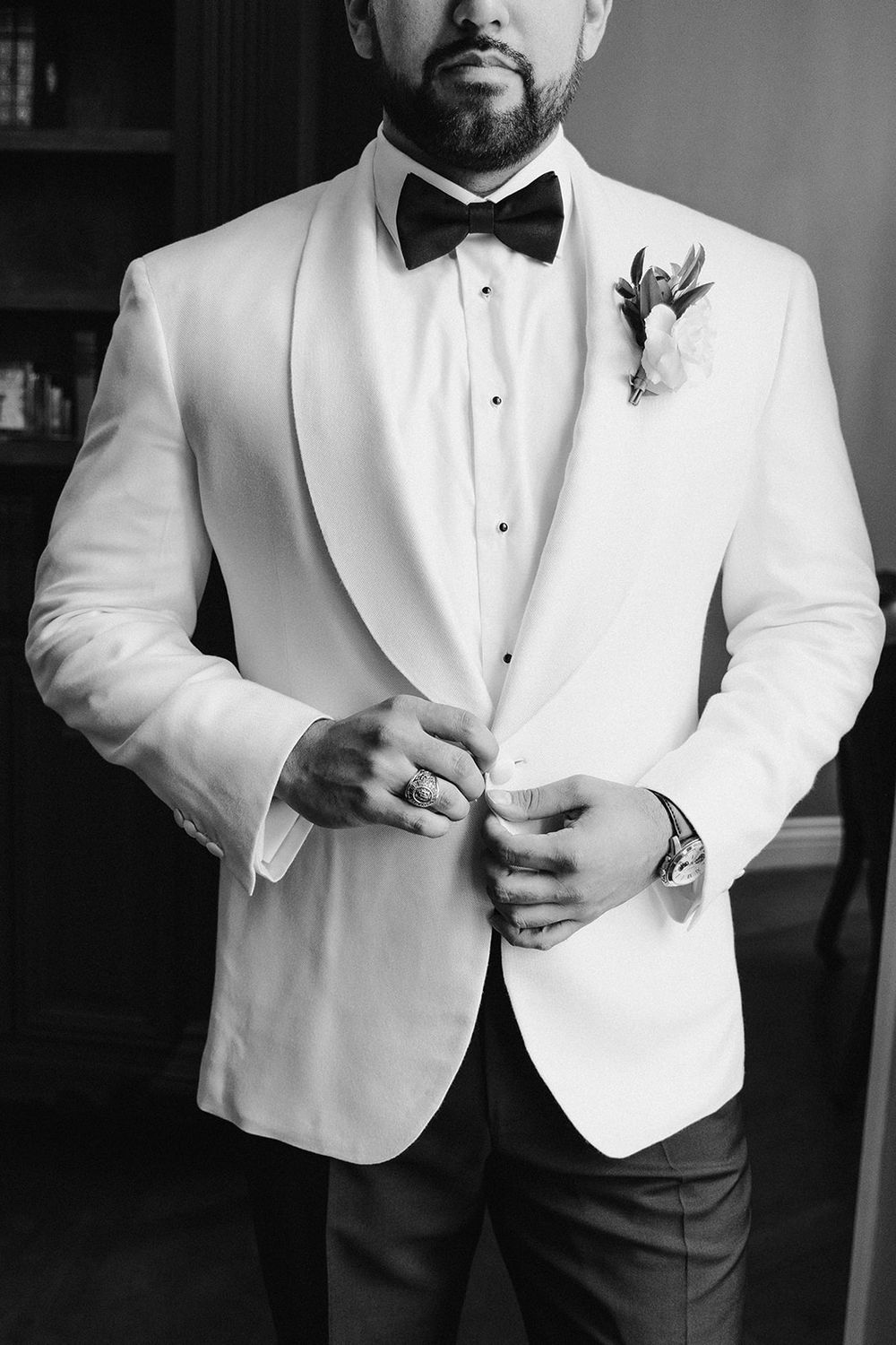 Groom in white tuxedo jacket with bow tie and boutonniere buttoning jacket, black and white wedding photo.