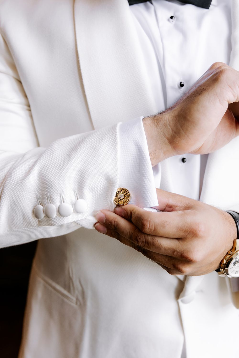A groom in a white tuxedo fastens a gold ornate cufflink on his dress shirt sleeve, wearing a luxury watch.