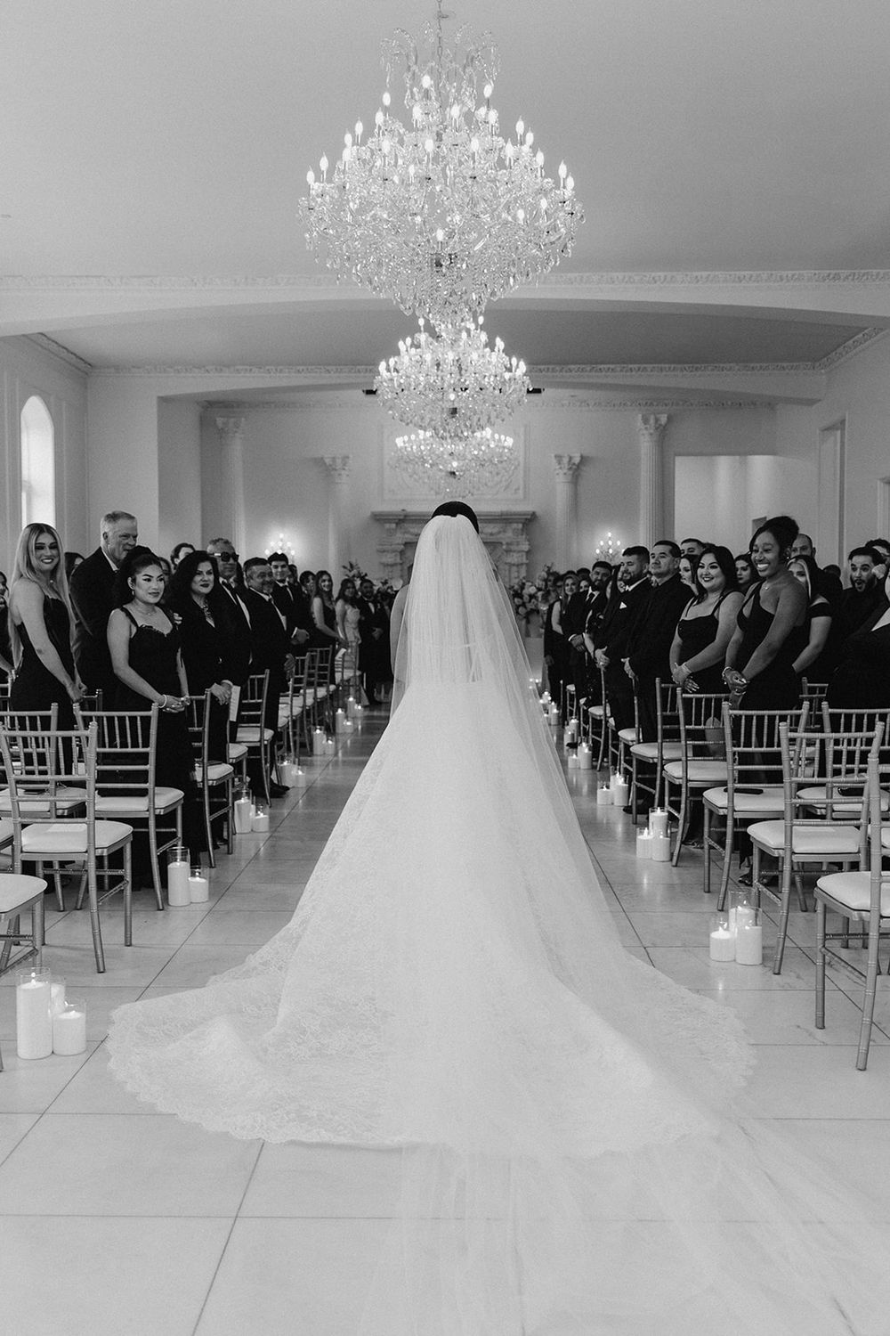 Black and white photo of a bride walking down the aisle in an elegant ballroom with a chandelier and guests seated.