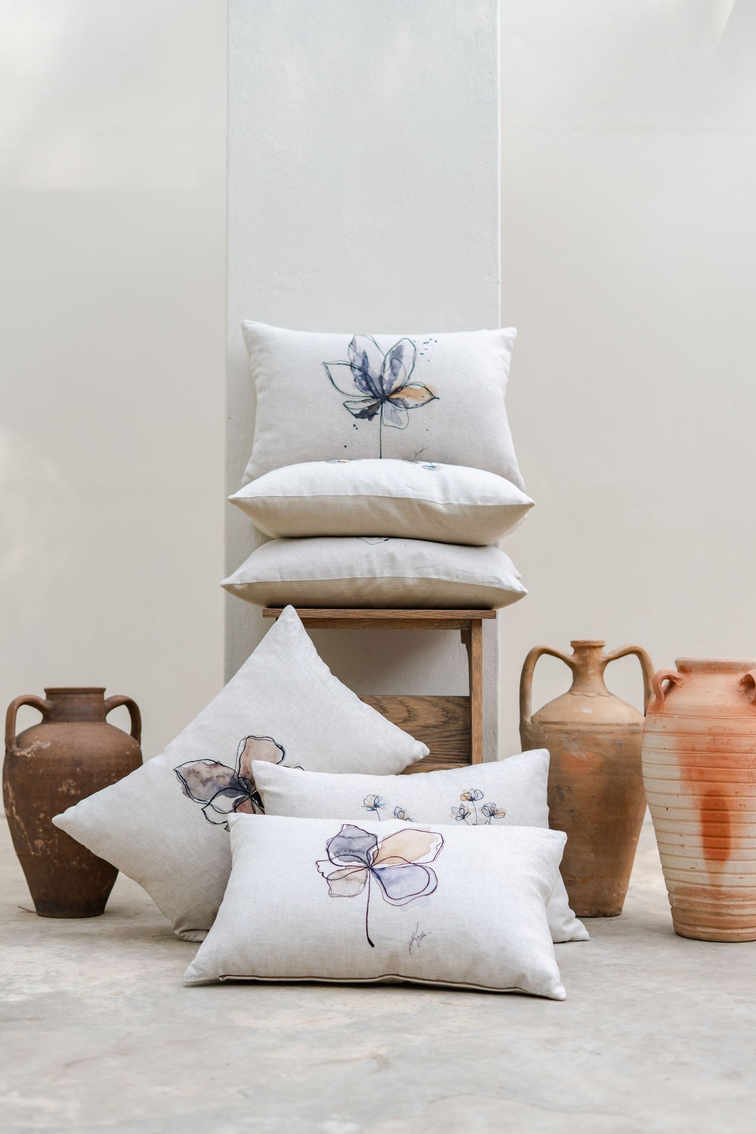 White linen pillows with hand-stitched watercolor floral prints styled with rustic terracotta vases on a neutral background.
