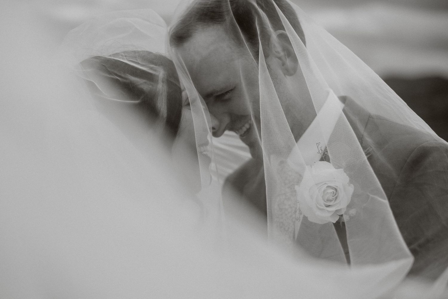 Black and white wedding photo of bride and groom sharing intimate moment under flowing veil, groom wearing rose boutonniere.