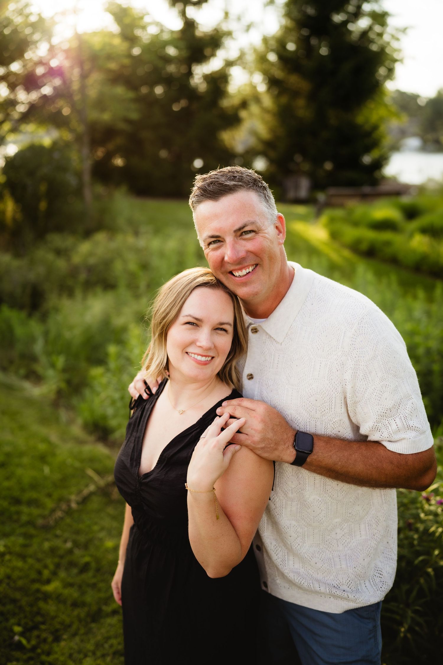 Smiling couple embracing outdoors in golden hour sunlight, woman in black dress and man in white shirt.