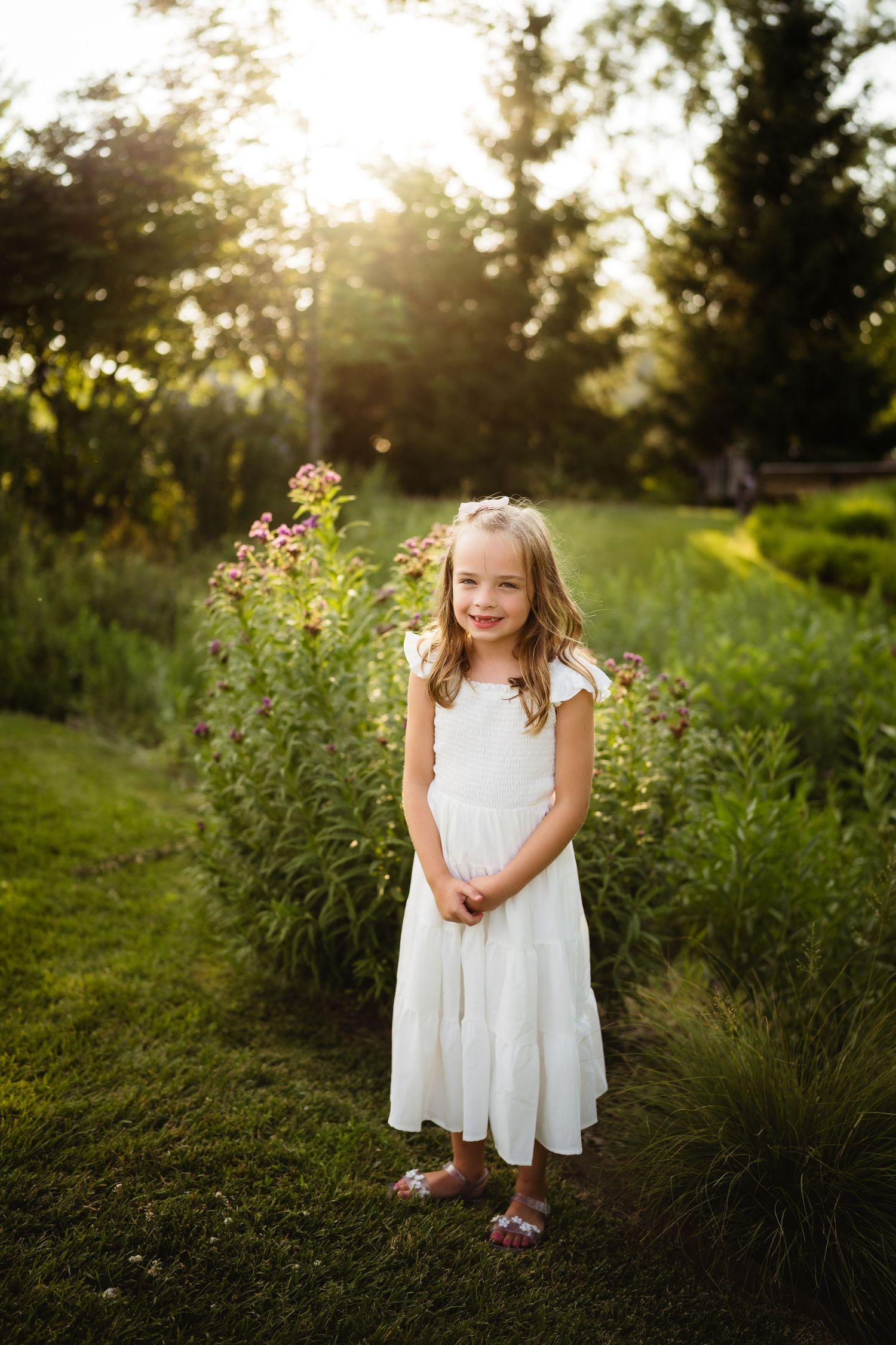 Young girl in white dress standing in sunlit garden with pink wildflowers and lush green trees in background.