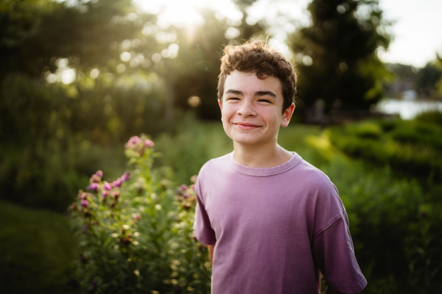 Smiling teenage boy in purple t-shirt posing outdoors in a lush green garden with pink flowers at golden hour.