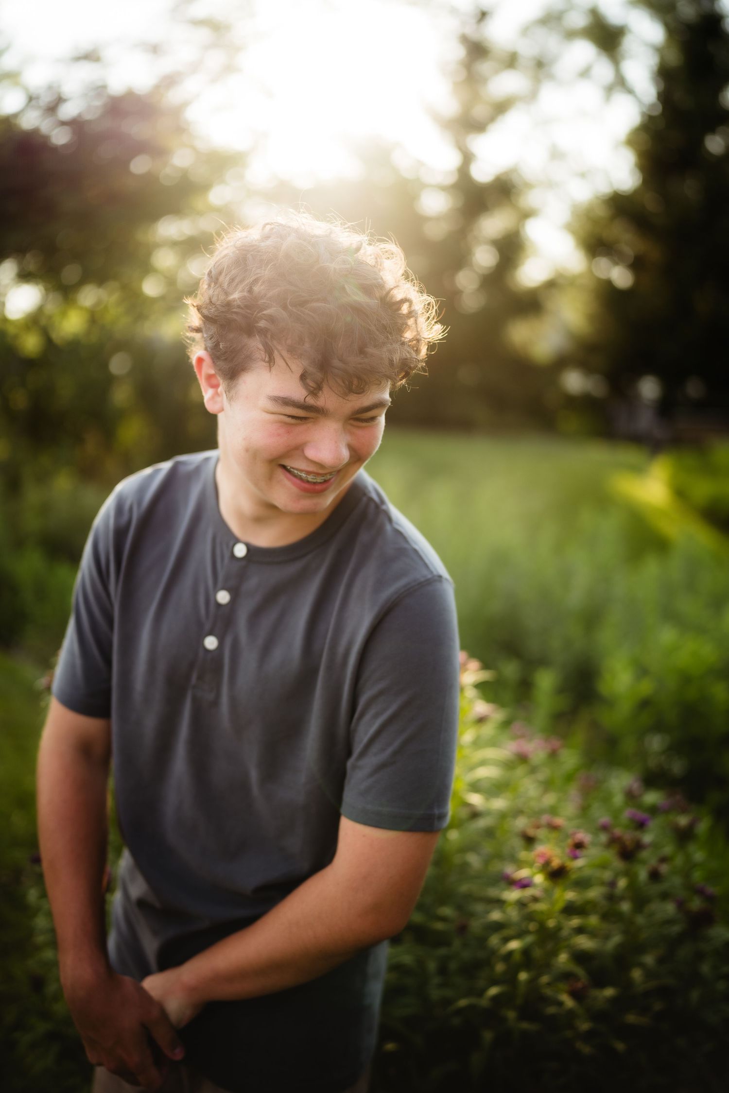 A smiling teenage boy with curly hair wearing a gray t-shirt stands outdoors in a sunlit garden.