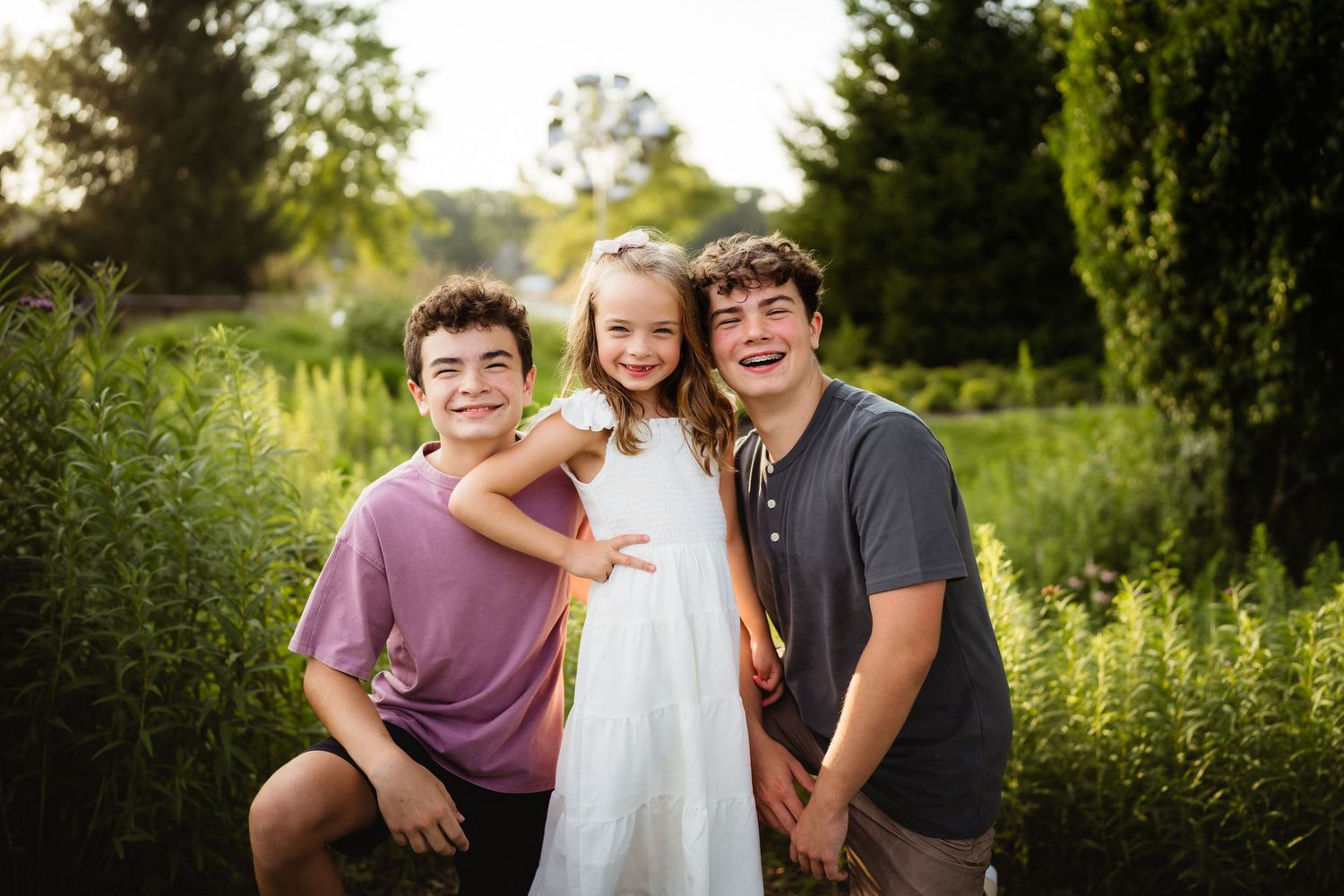 Three smiling children pose outdoors in a lush green garden, two boys and a girl in a white dress wearing a floral crown.