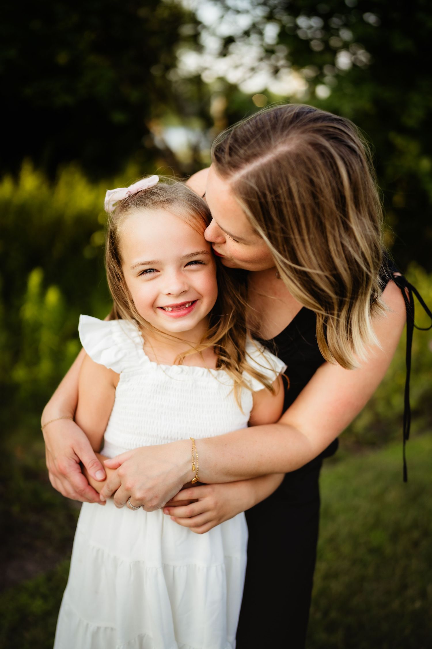 A woman in black dress hugs and kisses a smiling little girl in white dress outdoors in a green setting.