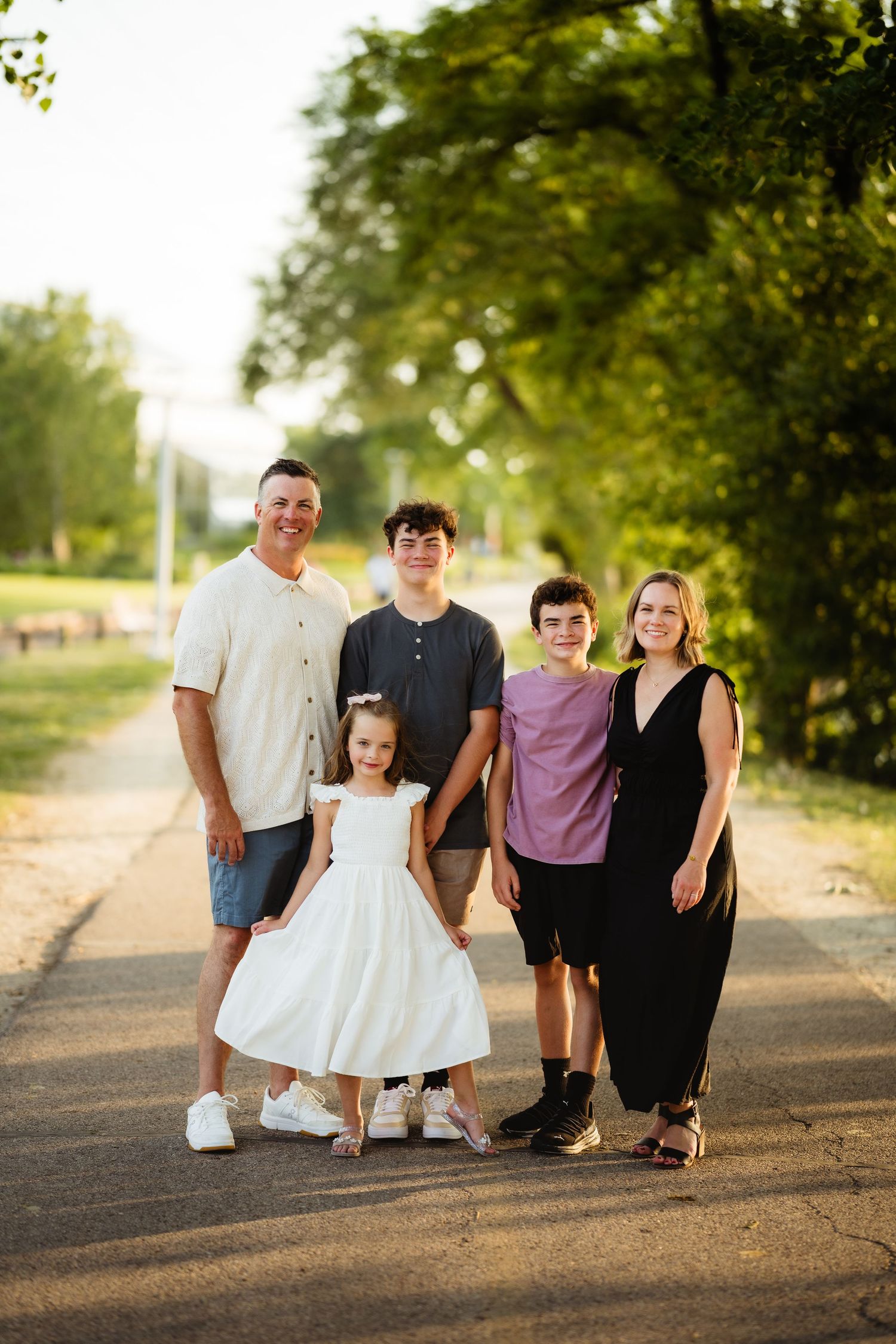 Family of five posing on a tree-lined path in golden hour sunlight, including two teens, a young girl in white dress.