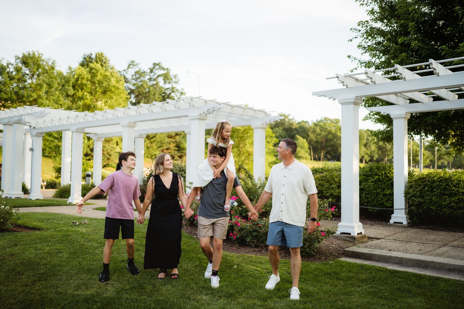 Family of five walking together under white pergola in lush green garden, smiling and holding hands on sunny day.