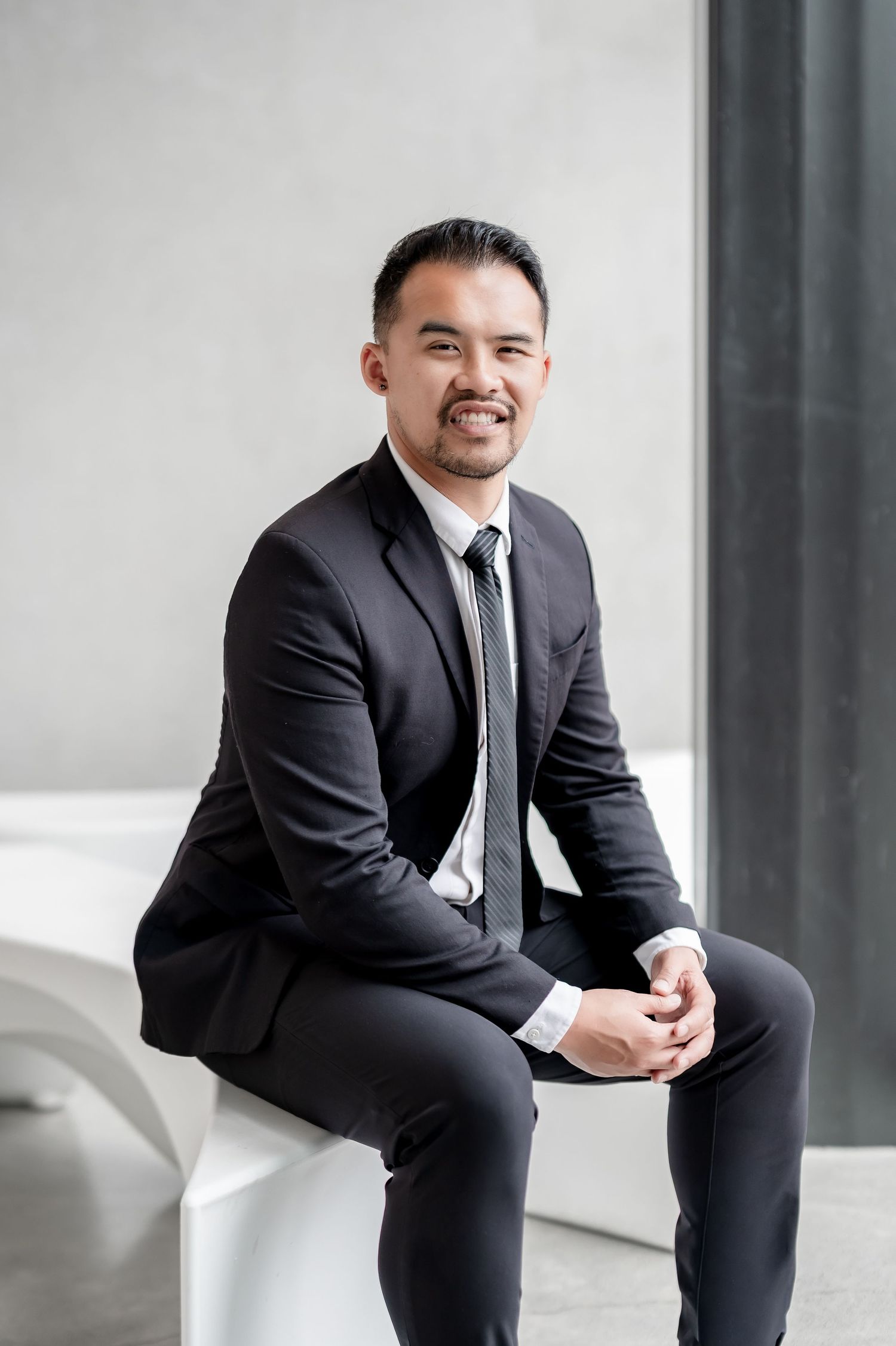 A professional Asian man in a black suit and tie sits smiling on a white bench in a modern interior setting.