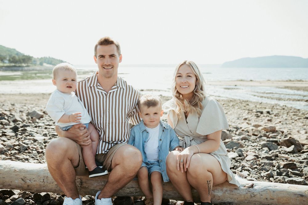 Family of four smiling on a rocky beach, parents holding young children, with ocean and mountains in background.