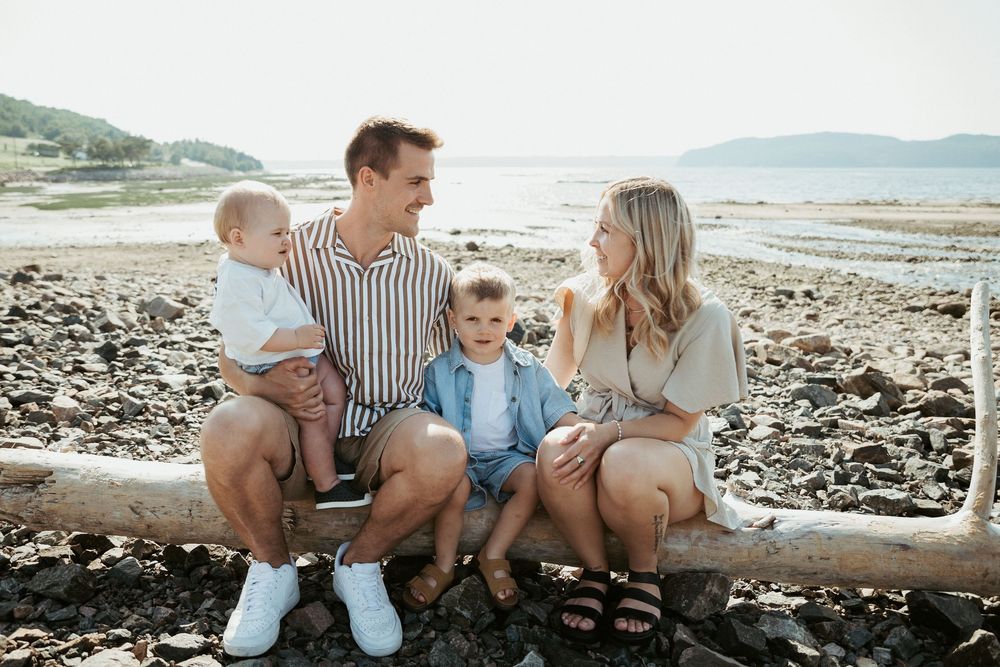 Family of four sitting on driftwood at a rocky beach, parents smiling at each other while holding two young children.