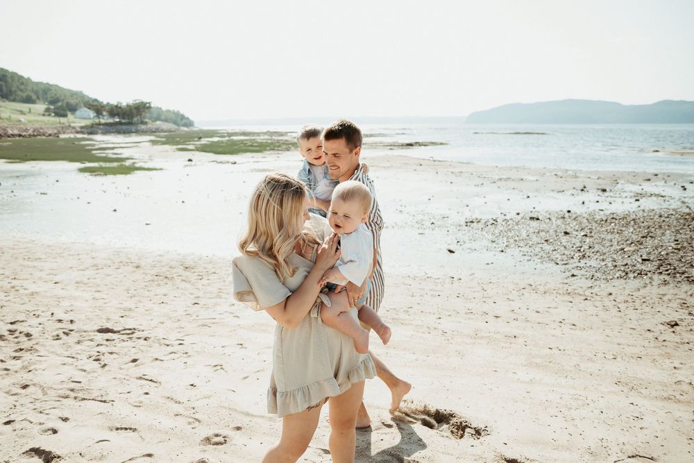 Family of four enjoying a sunny beach day, parents holding young children while walking along the sandy shoreline.