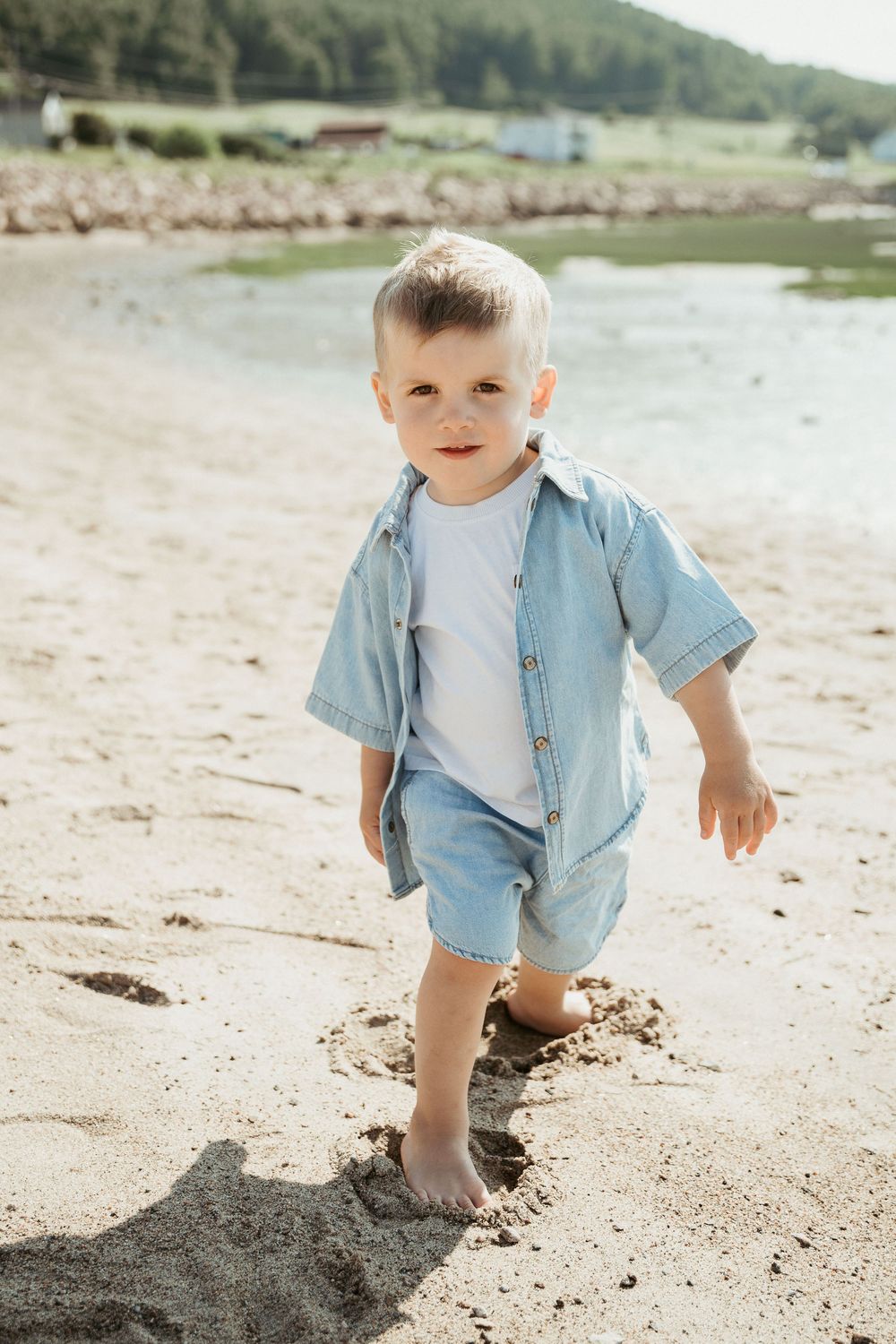 Toddler boy in denim shirt and shorts walking barefoot on sandy beach near calm water on sunny day.