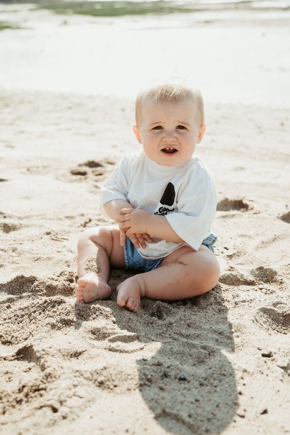 A happy baby sits barefoot in the sand at the beach, wearing a white tee and denim shorts on a sunny day.