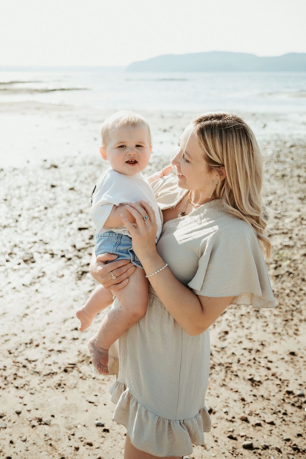 A blonde woman in a sage dress holds her smiling toddler on a sunny beach with mountains in the background.