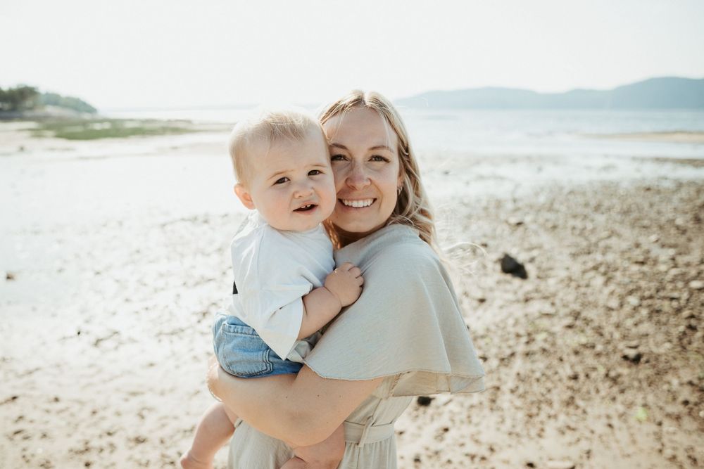 Smiling woman holding a happy baby on a sandy beach with calm water and mountains in the background.