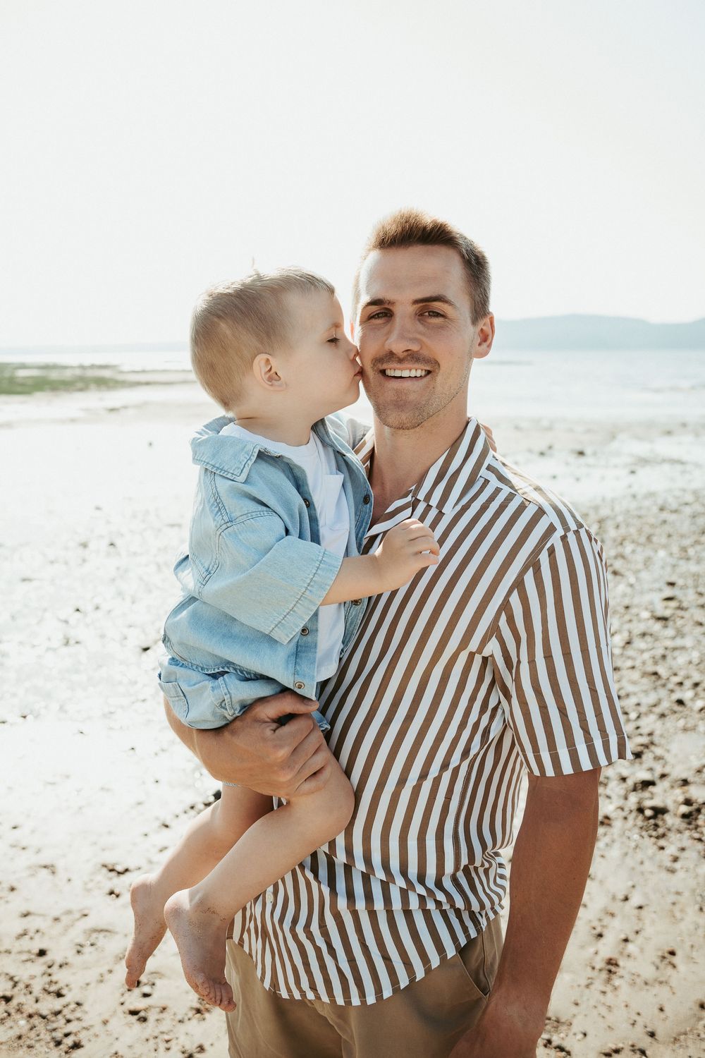 A smiling man holds a young child who kisses his cheek on a sunny beach.
