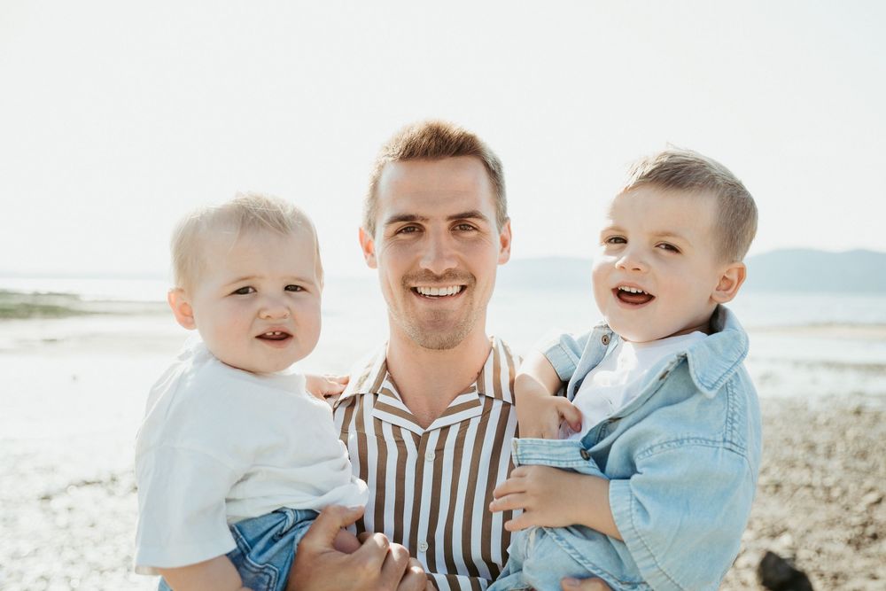A smiling man holds two happy toddlers on a sunny beach, enjoying a cheerful family moment outdoors.