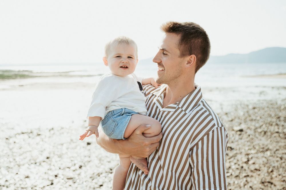 A smiling father holds his happy baby on a sunny beach, both wearing casual summer outfits near the ocean shore.