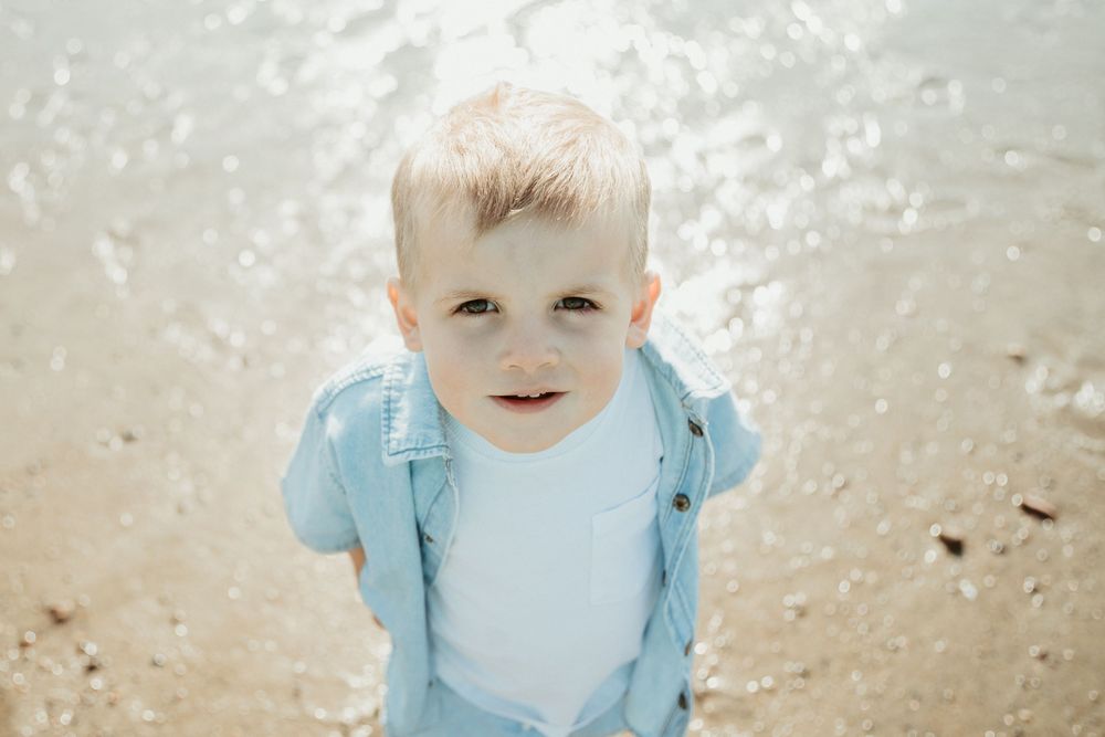Blonde toddler boy in blue denim jacket smiling at camera on sandy beach.