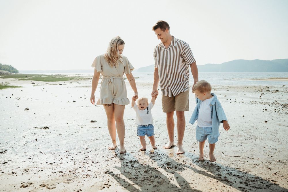 Family of four walking on a sandy beach, parents holding toddler hands, sunny day with mountains in background.