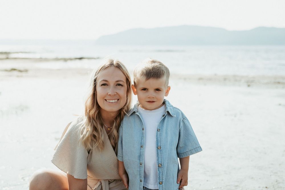 A smiling woman and young boy pose together on a sandy beach with mountains and calm water in the background.