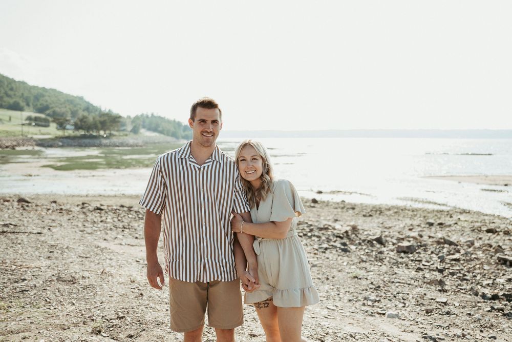 Couple smiling together on a rocky beach shoreline with calm water and green hills in the background.