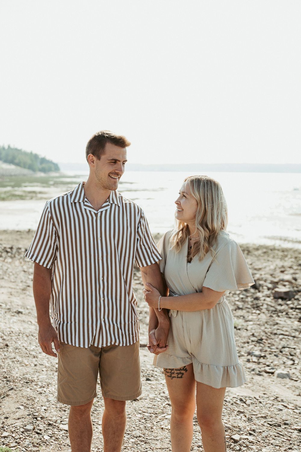 A smiling couple holds hands on a rocky beach, the man in a striped shirt and the woman in a beige romper.