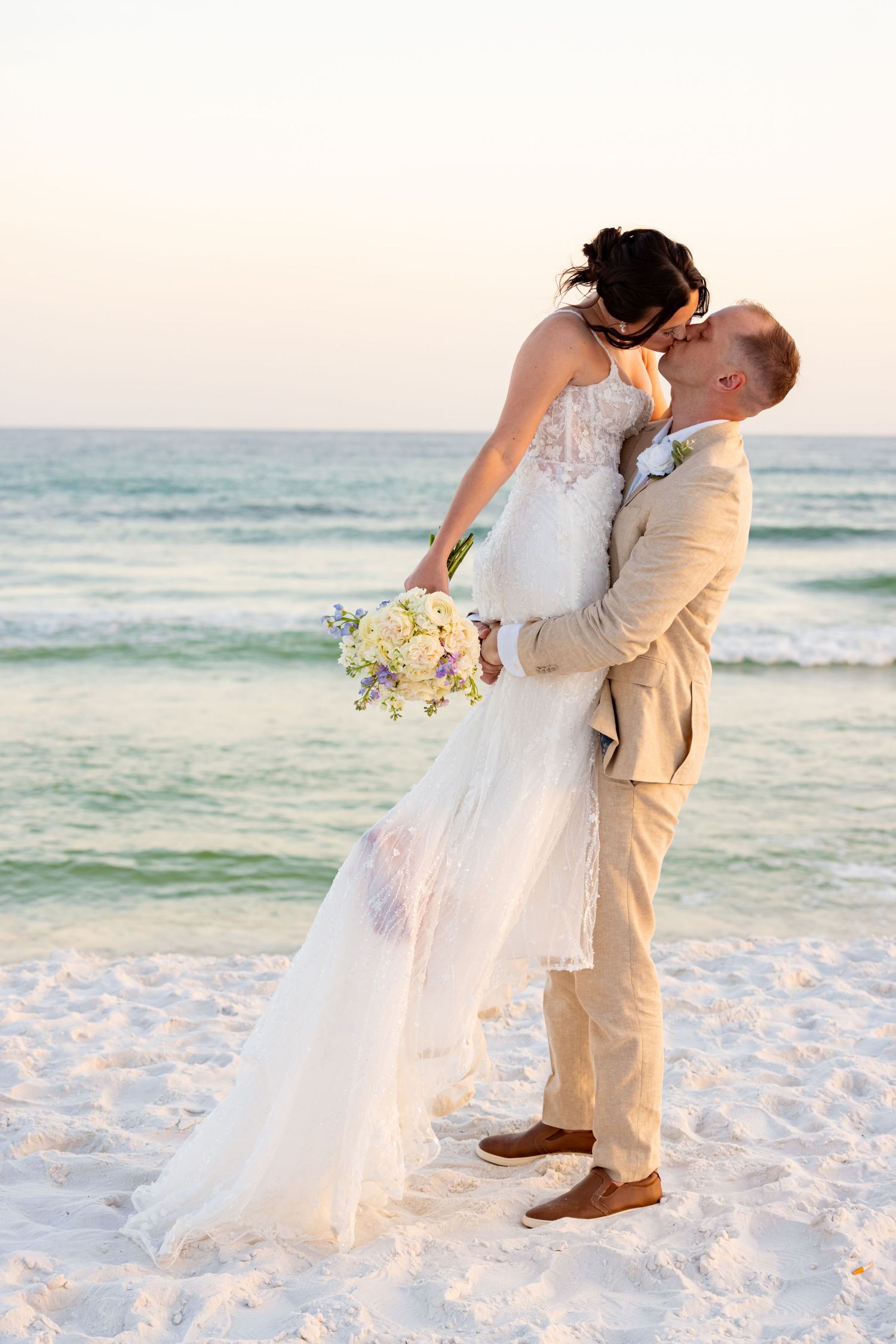 Bride and groom share a romantic beach kiss at sunset, bride holding a pastel bouquet in a white gown, groom in tan suit.