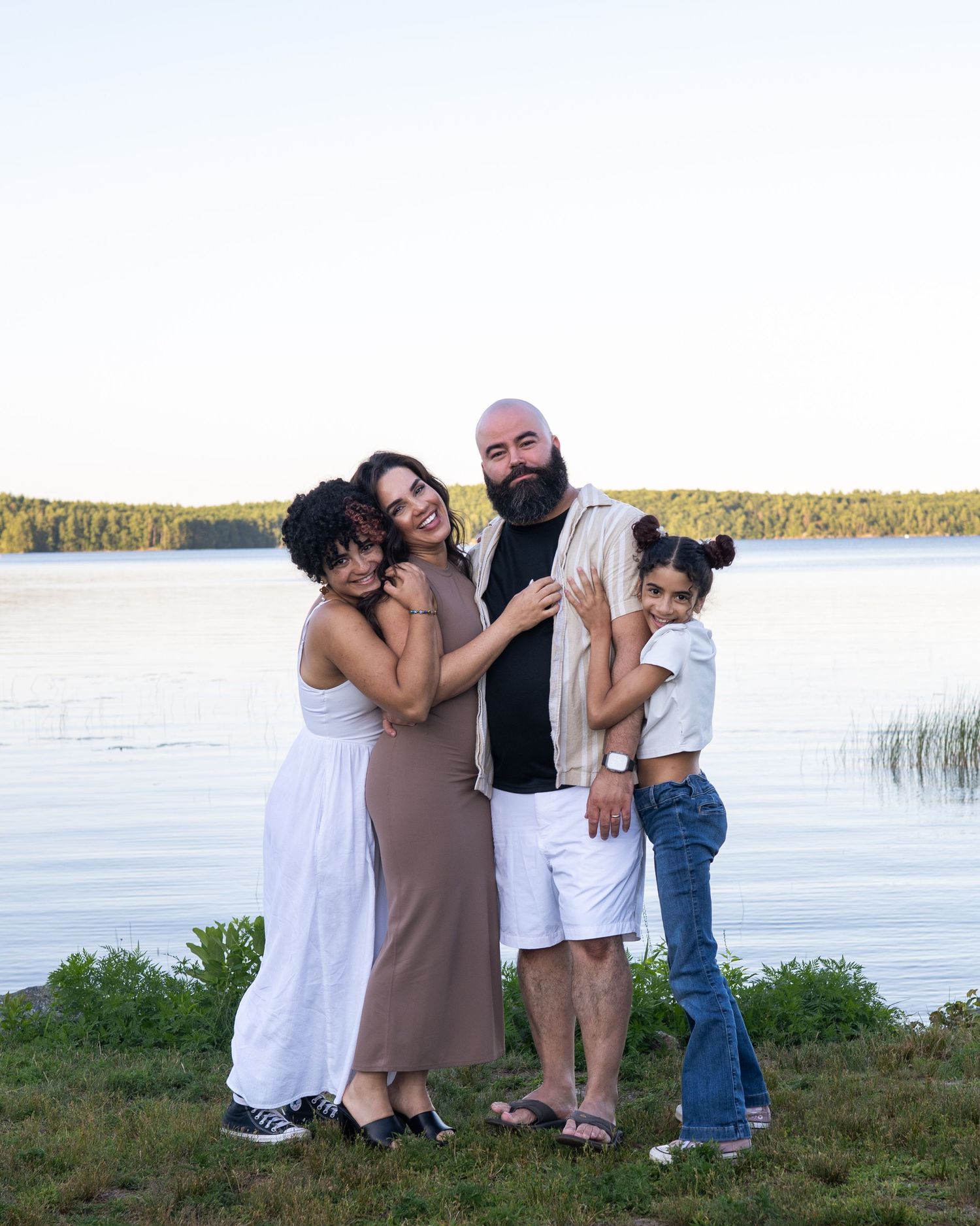 Family of four posing together by a serene lake, smiling warmly in casual summer outfits at dusk.