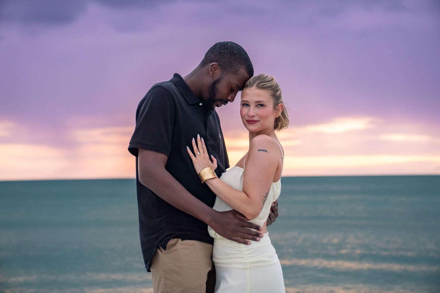 Couple embracing on a beach at sunset with purple sky and turquoise ocean, woman in white dress smiling at camera.