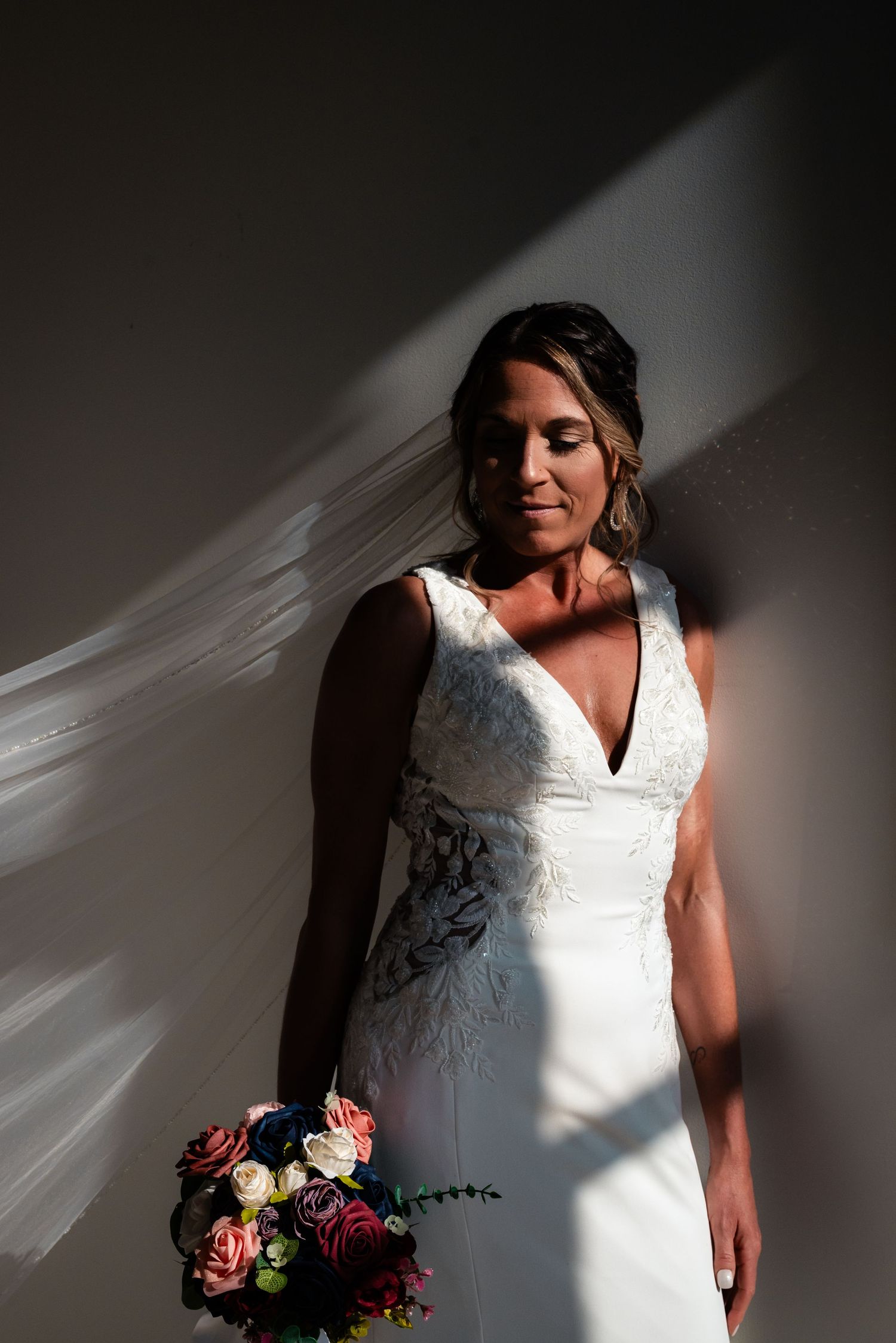 Bride in white lace gown holding colorful bouquet, standing against wall in dramatic light and shadow.