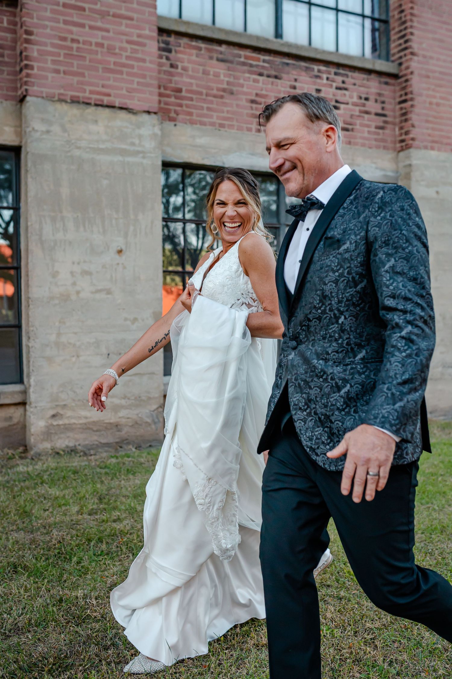 Bride in white lace gown and groom in dark floral tuxedo jacket laughing together outside brick building.