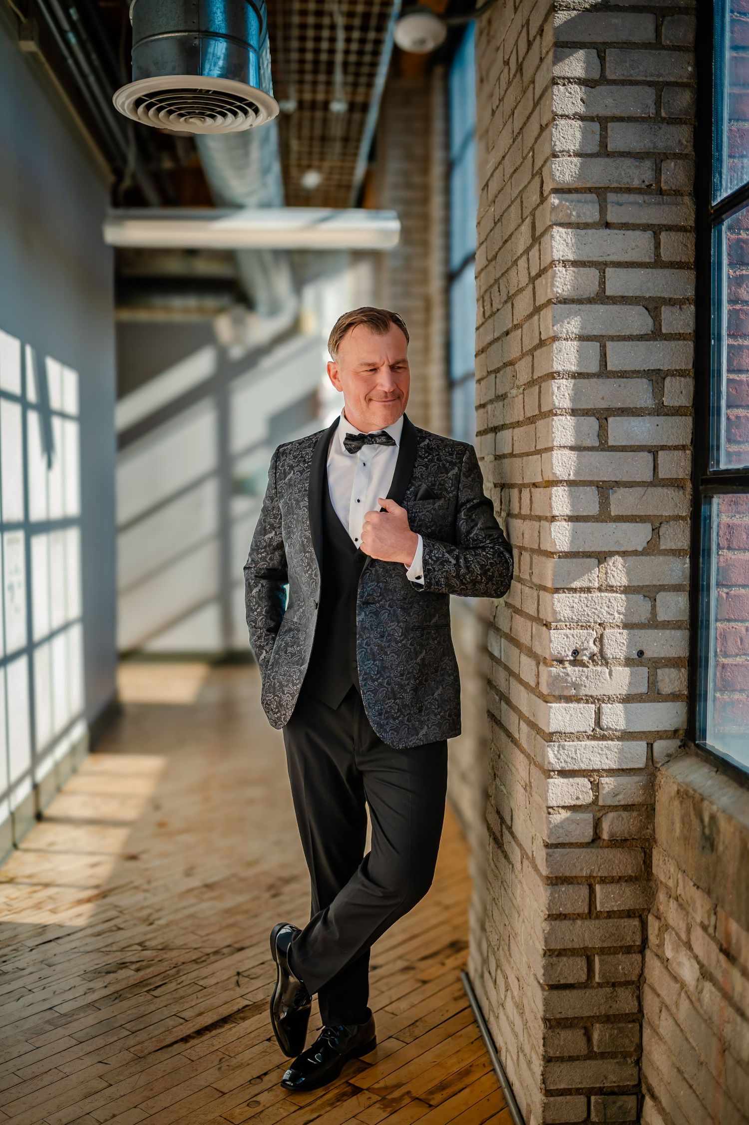 A dapper groom in a floral tuxedo jacket and bow tie leans against a brick wall in a sunlit industrial venue.