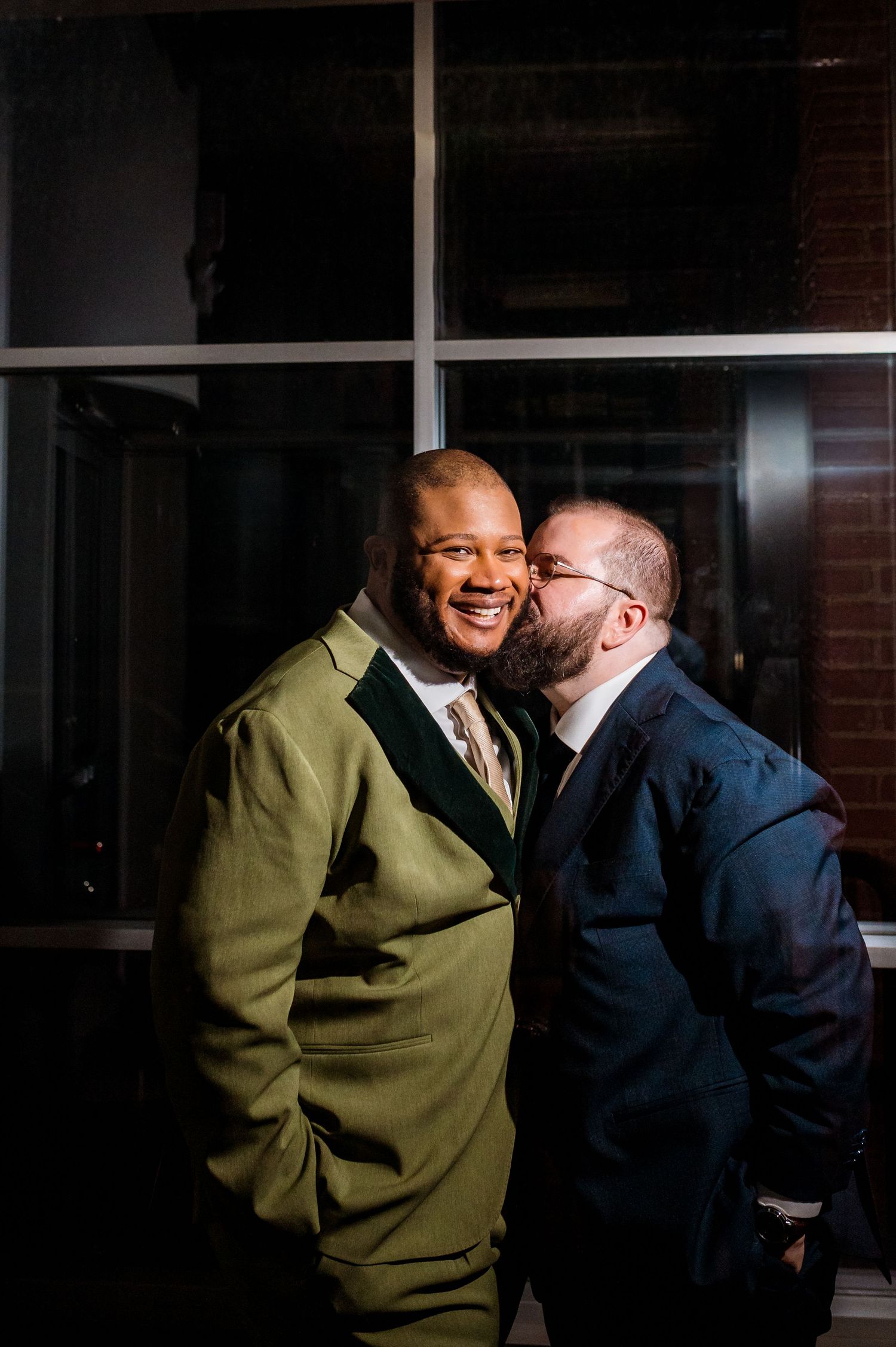 Two grooms share a tender moment, one in olive suit smiling as the other in navy suit gives a cheek kiss.
