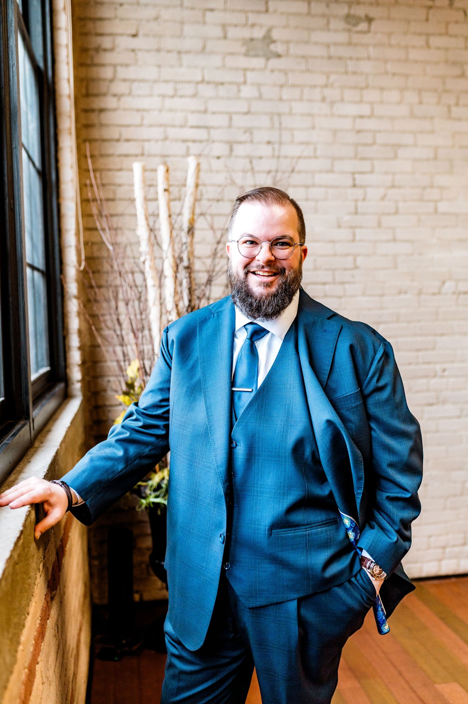 Smiling bearded man in blue three-piece suit and glasses posing near window against white brick wall.