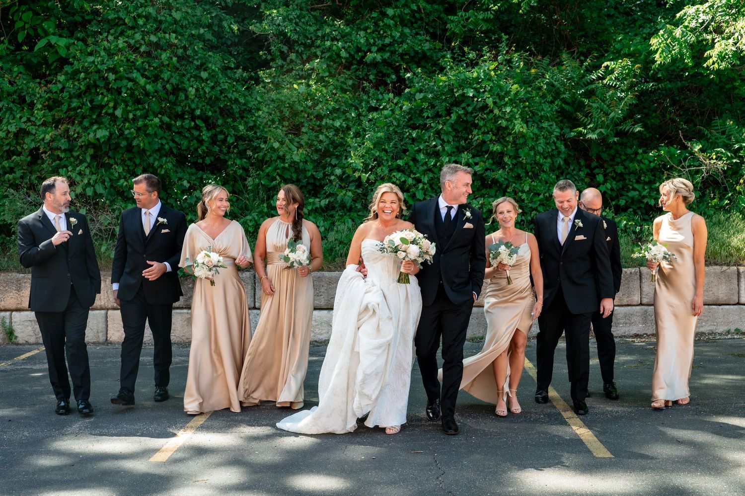 Wedding party in formal attire walks outdoors, bridesmaids in champagne gowns, groomsmen in black tuxedos, bride in white gown.