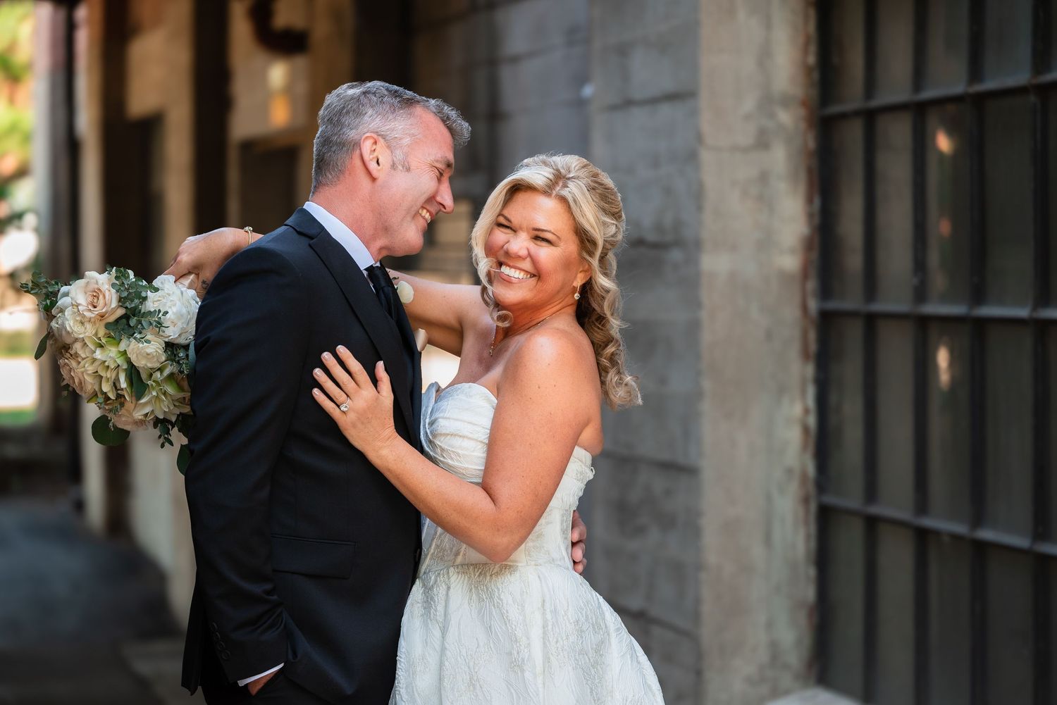 Bride and groom embrace outdoors, smiling joyfully, bride holding white bouquet in elegant strapless gown.