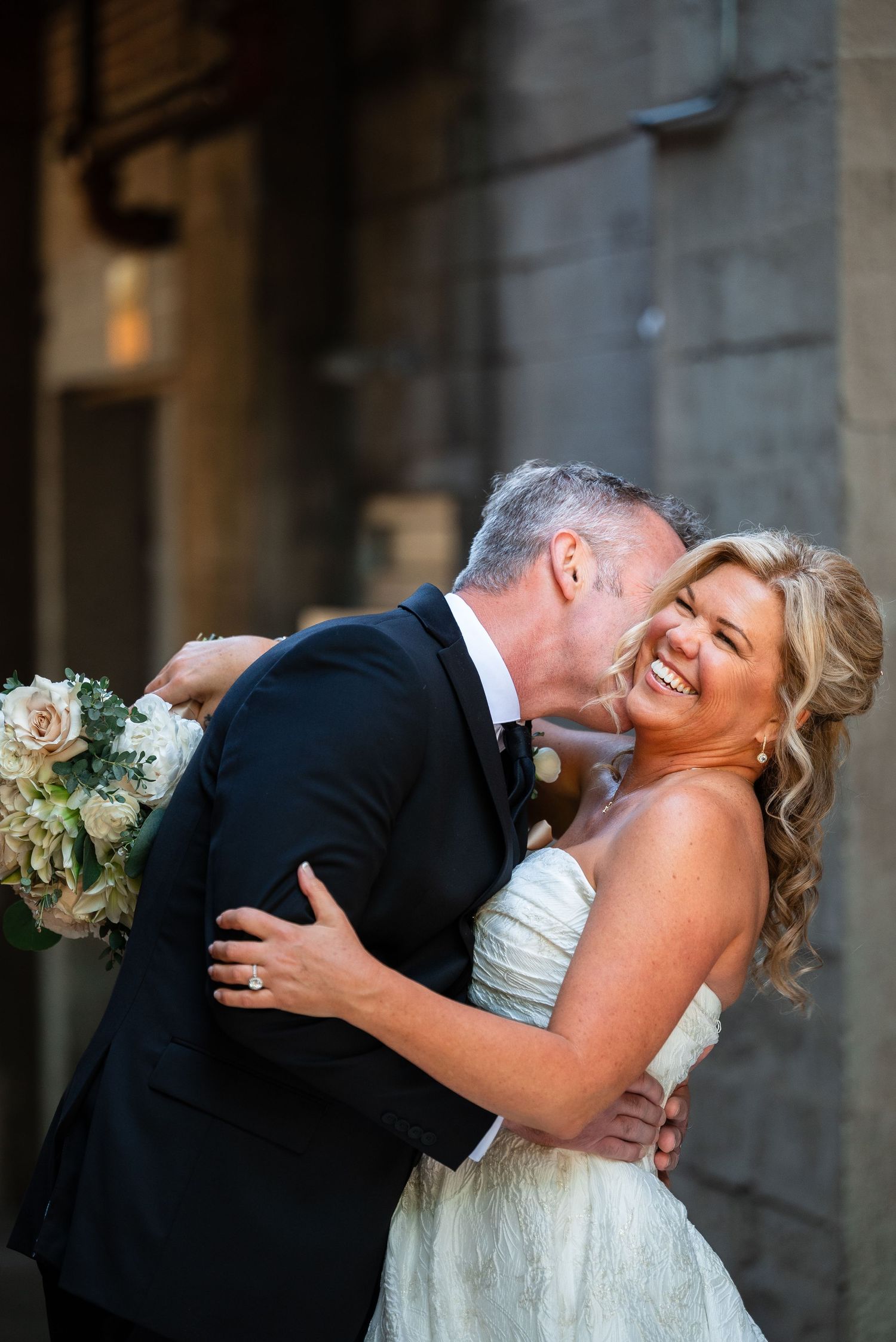 Bride and groom share a joyful embrace, groom kissing bride's cheek as she laughs, holding white floral bouquet.